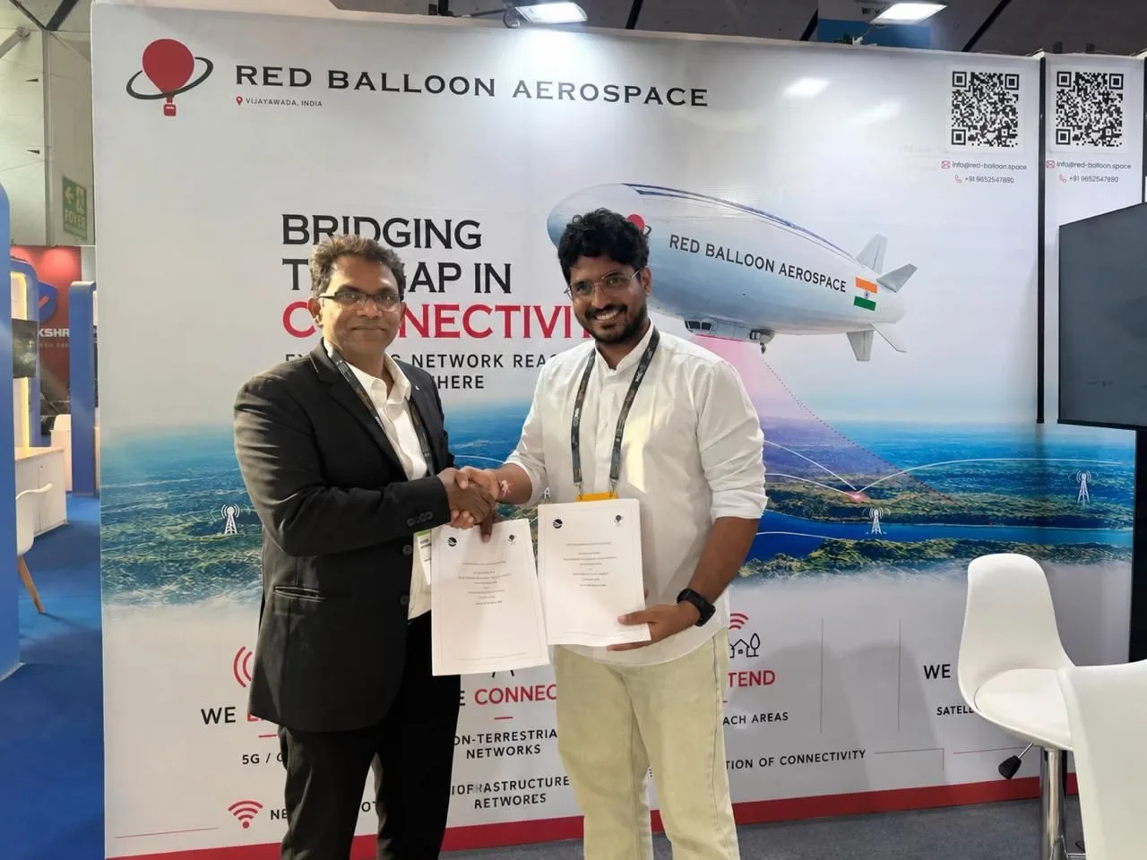 Two people shaking hands and holding signed documents in front of the Red Balloon Aerospace stand